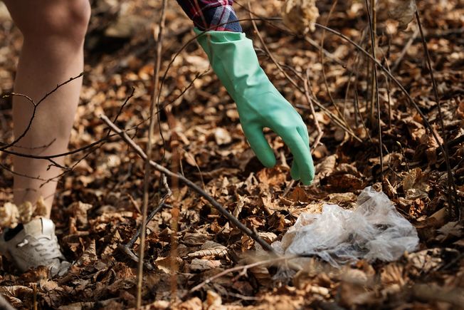 Plogging, den fysiske øvelsen der søppelplukking og jogging kombineres, til glede for både utøver og planet. En hånd med gummihanske strekker seg etter et stykke plast blant høstløvene.