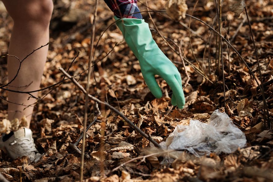 Plogging, den fysiske øvelsen der søppelplukking og jogging kombineres, til glede for både utøver og planet. En hånd med gummihanske strekker seg etter et stykke plast blant høstløvene.