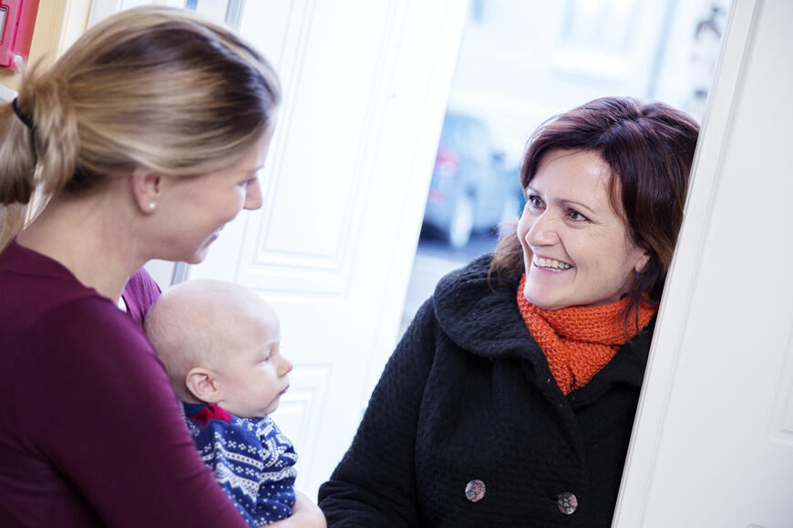 Bilde av en frivillig familiekontakt som kommer på besøk til en dame med en baby i armene.