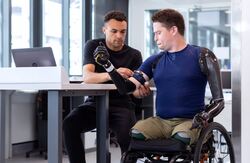 Robotarmer og -bein på mann i rullestol. Mann ved siden av hjelper ham å sette på.
