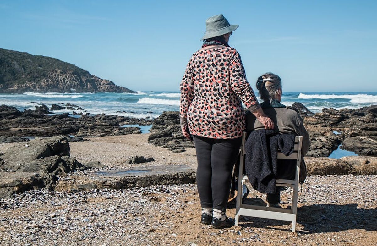 To eldre kvinner med ryggen til ved en strand. En sitter på en stol. Den stående har hatt.