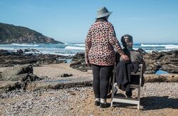 To eldre kvinner med ryggen til ved en strand. En sitter på en stol. Den stående har hatt.