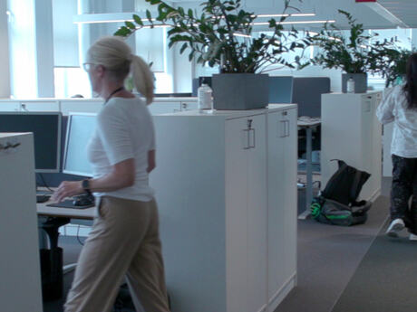 To kvinner i et kontorlandskap, der vi ser noen planter og en del arkivskap, et par PCer