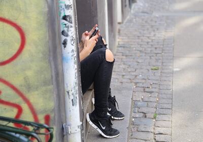 Tenåring sitter ved vegen, ser kun bein med sorte jeans og joggesko, og hender som holder mobiltelefon.