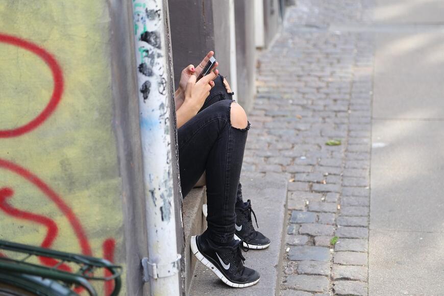 Tenåring sitter ved vegen, ser kun bein med sorte jeans og joggesko, og hender som holder mobiltelefon.