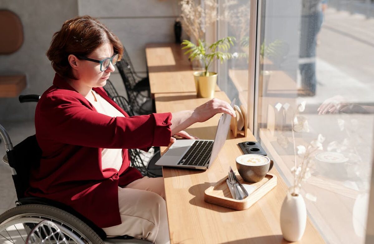 Kvinne i rullestol sitter med PCen ved et vindusbord. En asjett med noe mat og en kaffe.