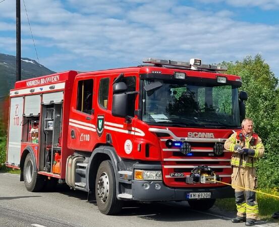 Bildet viser en stor brannbil med en brannmann foran bilen. Det er fint vær.