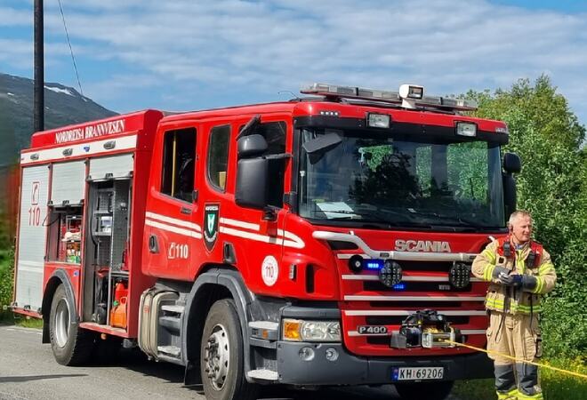 Bildet viser en stor brannbil med en brannmann foran bilen. Det er fint vær.