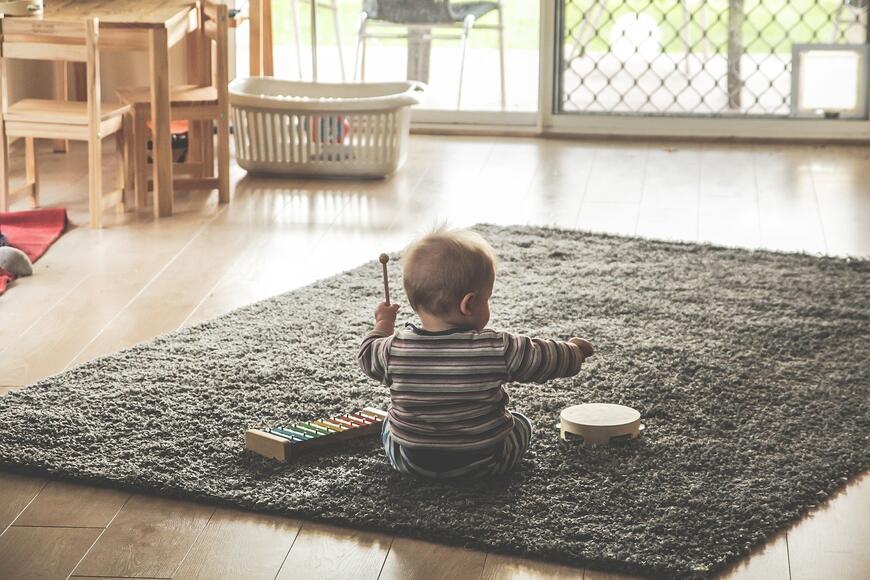 En baby sitter på et teppe på gulvet og spiller på en liten tromme og en xylofon. Det er sett bakfra.