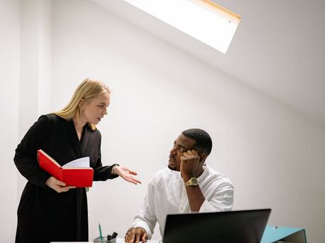 hvit kvinne står og gir tilbakemelding til en mørk mann som sitter