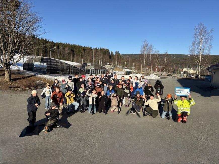 Barn som står på en skoleplass, noen av barna holder sparkesykler.
