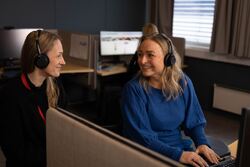 To kvinner i NAV kontaktsenter, begge med hodetelefoner. De ser på hverandre og smiler