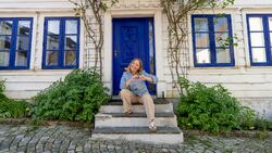 Smilende kvinne på trapp foran et idyllisk hus i hvitt og blått.