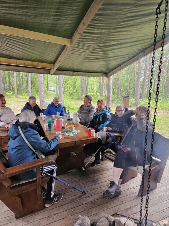 På bildet sitter det glade mennesker rundt et bord ute i skogen. De flirer og har det artig.