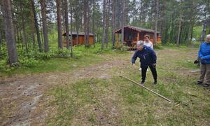 På bildet ser vi en person som skal kaste en hestesko. Bak henne er det to spente deltakere som følger nøye med på hvor langt personen kaster. I bakgrunnen ser vi furuskog og to tømmerhytter.