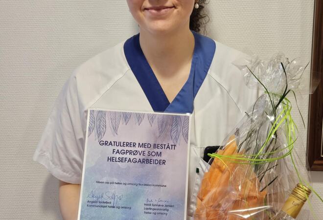 Bildet viser en meget fornøyd dame som har bestått fagprøven som helsefagarbeider. Hun smiler bredt mens hun holder et diplom og en gave.