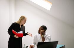 En kvinne med langt lyst hår og mørk kjole holder en bok med rødt omslag i hendene, og står bøyd litt over en mørk mann med hvit skjorte som sitter ved en PC.