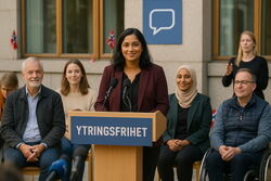 Talerstol, kvinner og menn, kvinne med mørkt hår taler, norske flagg rundt