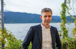 Mann i dress ser inn i kameraet, natur i bakgrunnen