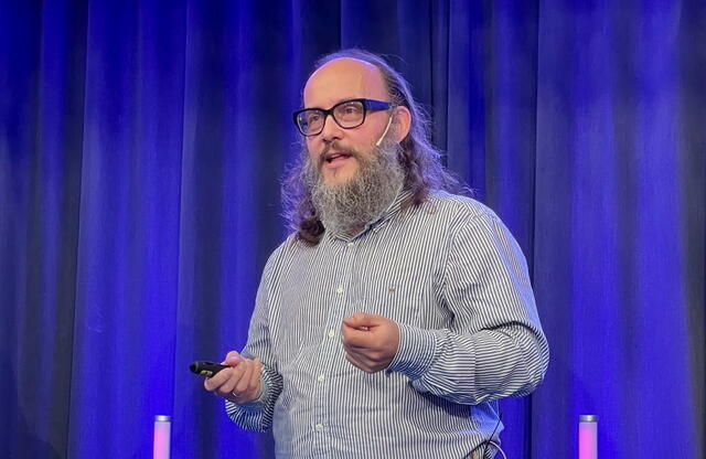 – Vi blir målt på tillit, og da må språket vårt gjøre én ting: hjelpe folk å handle riktig, sa Lars Rønn. Mann med skjegg og langt hår på scenen