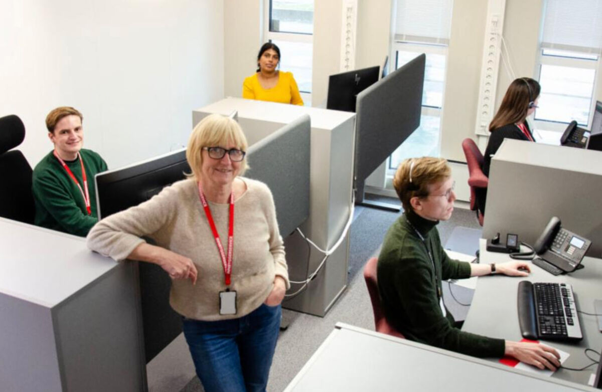 Lillian Røthe, kvinne, står avslappet i et kontorlandskap med sittende personer