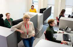 Lillian Røthe, kvinne, står avslappet i et kontorlandskap med sittende personer