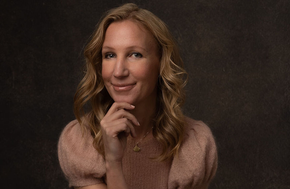 Kvinne med langt lyst hår, en hånd under haken, ser tenksomt mot kamera