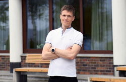 Mann i T-skjorte, ungdommelig, guttaktig, armene i kors, utendørs, bygning i bakgrunnen med glassfasade og hvite søyler.