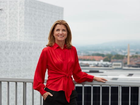 Nina Schanke Funnemark står utendørs, muligens på et tak der hun holder i et rekkeverk med en hånd. Hun smiler, og har på en rød løsthengende bluse. Håret er mellomblondt og langt. I bakgrunnen er det et bymiljø.