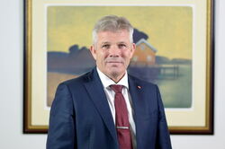 Bjørnar Skjæran med et maleri i bakgrunnen. Han har blå dress, burgunder slips og hvit skjorte. Grå i håret.