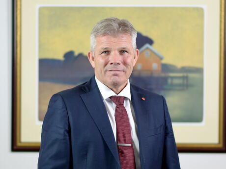 Bjørnar Skjæran med et maleri i bakgrunnen. Han har blå dress, burgunder slips og hvit skjorte. Grå i håret.