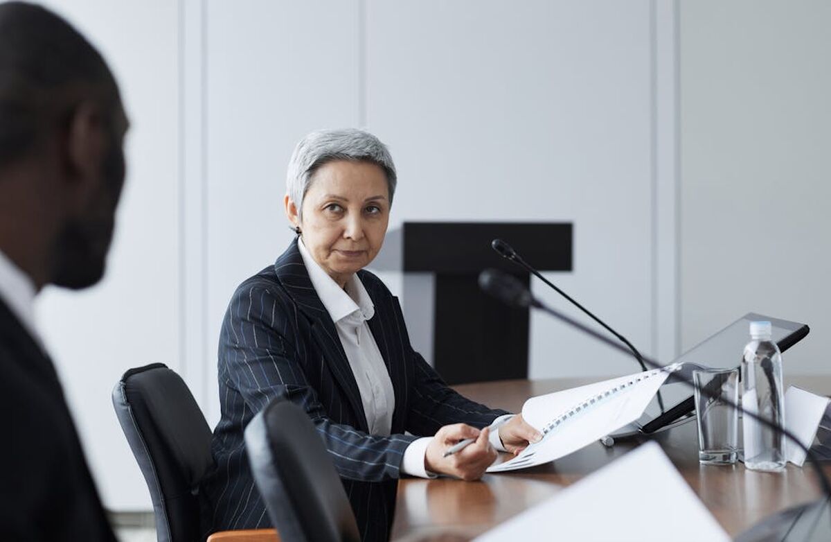 Kvinne med kort grått hår og mørk stripete jakke sitter ved et bord. I forgrunnen en utydelig farget person som vi bare ser bakhodet på.