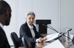 Kvinne med kort grått hår og mørk stripete jakke sitter ved et bord. I forgrunnen en utydelig farget person som vi bare ser bakhodet på.