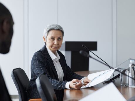 Kvinne med kort grått hår og mørk stripete jakke sitter ved et bord. I forgrunnen en utydelig farget person som vi bare ser bakhodet på.