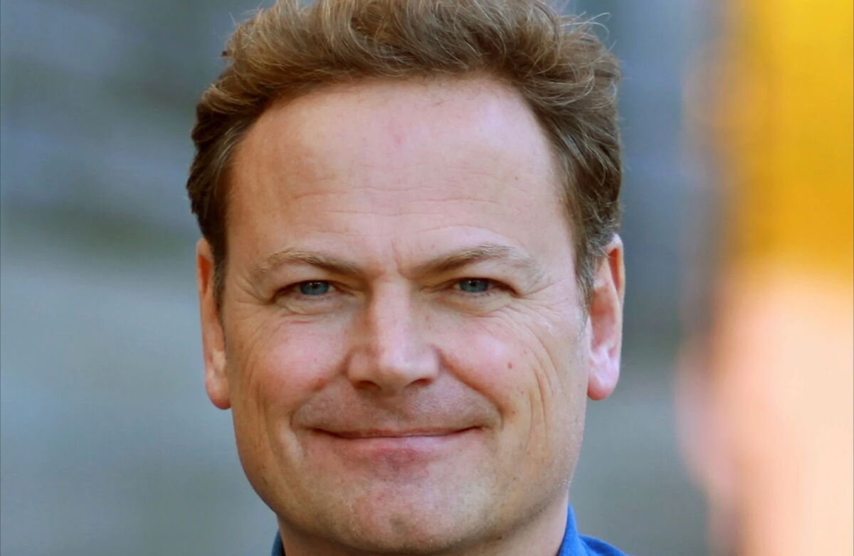 Mann med kortklipt brunt hår ser alvorlig inn i kameraet. Han har blå skjorte. Bakgrunnen er diffus.