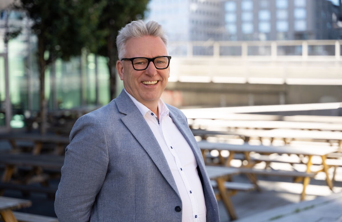 Smilende middelaldrende mann med lys dressjakke og hvit skjorte. Han står utendørs i et bymiljø.