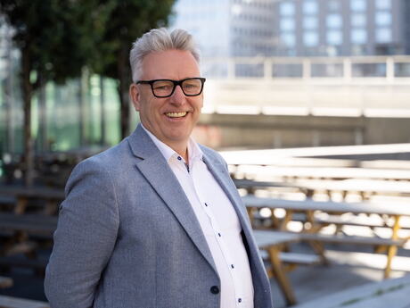 Smilende middelaldrende mann med lys dressjakke og hvit skjorte. Han står utendørs i et bymiljø.