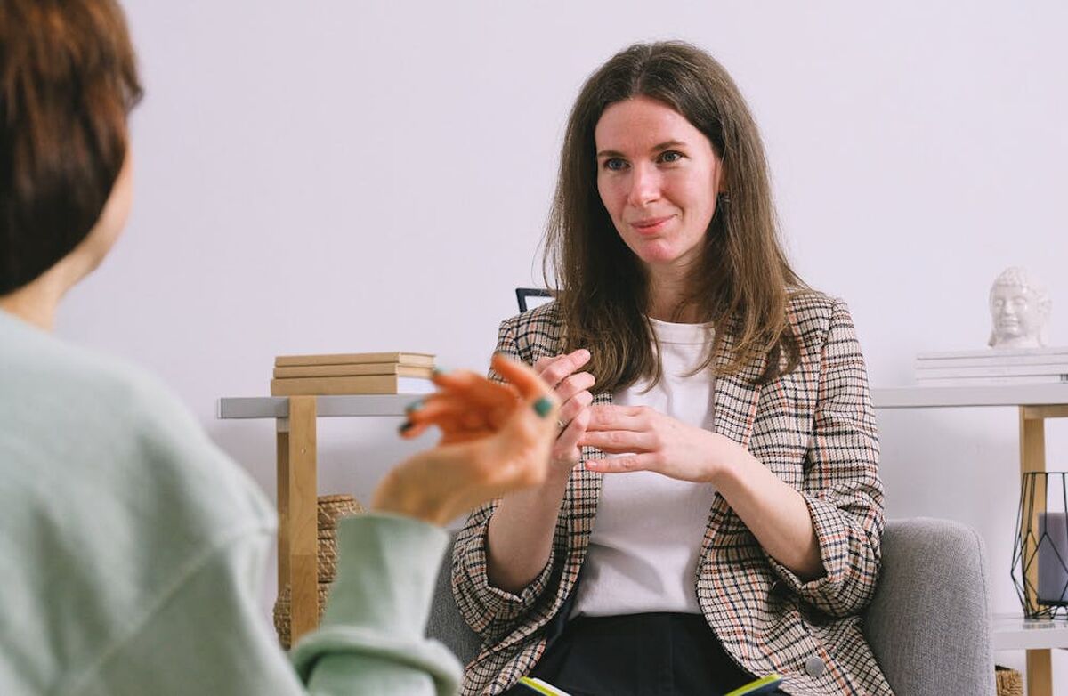 To kvinner sitter i et rom. Den nærmeste ser vi bakfra. Den andre ser empatisk ut.
