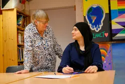 En eldre dame som står snakker med en yngre kvinne som sitter.