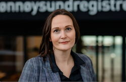 En kvinne med langt halvmørkt hår ser alvorlig inn i kameraet. I bakgrunnen ser vi en en uklar husfasade der vi ser deler av teksten