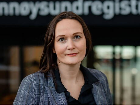 En kvinne med langt halvmørkt hår ser alvorlig inn i kameraet. I bakgrunnen ser vi en en uklar husfasade der vi ser deler av teksten