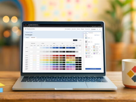 En åpen PC-skjerm på et trebord viser designsystemet. På bordet er det postit-lapper i forskjellige farger på venstre side og en kaffekopp på høyre side.