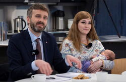 Middelaldrende mann i dress sitter ved siden av en ung kvinne med en baby i armene.