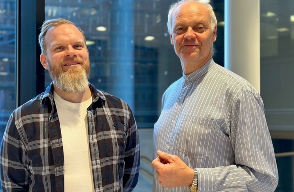 Bildet viser to menn som står side om side innendørs, foran store vinduer med et moderne kontorbygg i bakgrunnen.  * Mannen til venstre har kort hår og et lyst, fyldig skjegg. Han smiler og har på seg en rutete flanellskjorte over en hvit t-skjorte. *