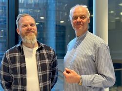 Bildet viser to menn som står side om side innendørs, foran store vinduer med et moderne kontorbygg i bakgrunnen.  * Mannen til venstre har kort hår og et lyst, fyldig skjegg. Han smiler og har på seg en rutete flanellskjorte over en hvit t-skjorte. *