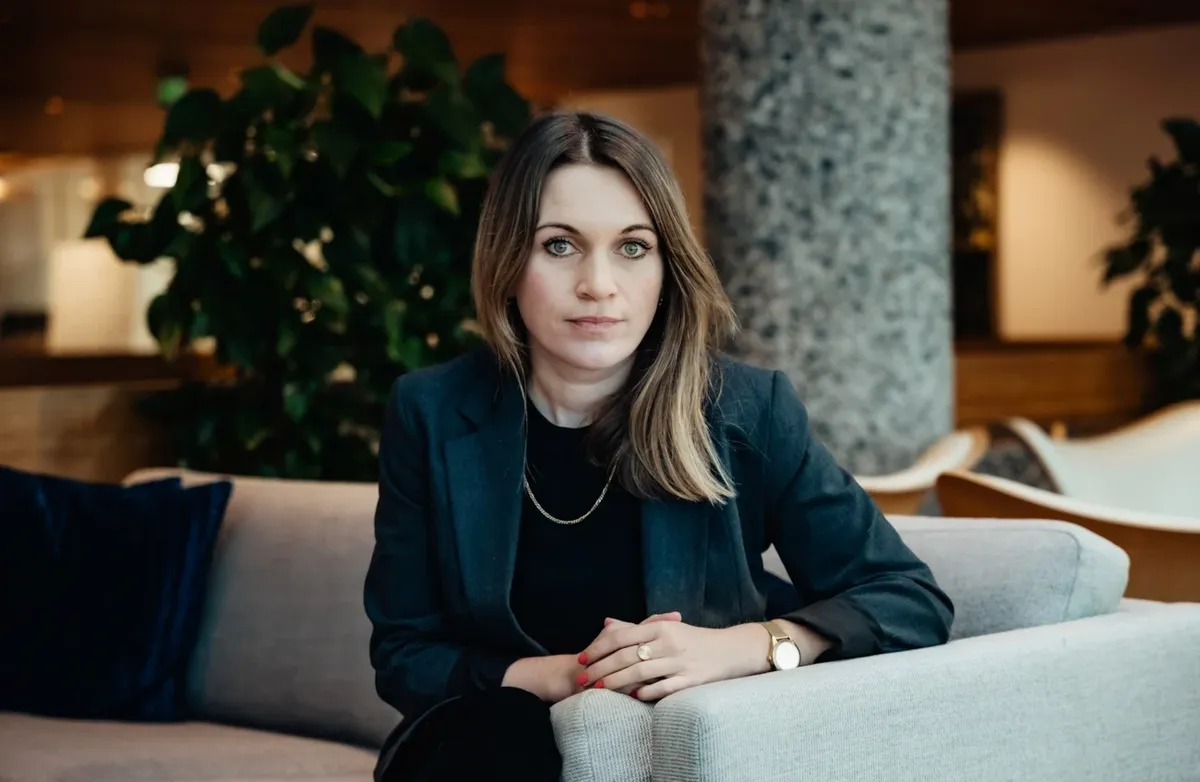 Bildet viser en kvinne som sitter i en lys sofa i et moderne innendørsmiljø, trolig et kontorbygg eller en hotellobby. Hun sitter vendt rett mot kameraet med hendene samlet foran seg på armlenet. Kroppsspråket er rolig og kontrollert, og hun har et di