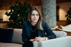 Bildet viser en kvinne som sitter i en lys sofa i et moderne innendørsmiljø, trolig et kontorbygg eller en hotellobby. Hun sitter vendt rett mot kameraet med hendene samlet foran seg på armlenet. Kroppsspråket er rolig og kontrollert, og hun har et di