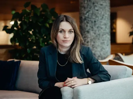 Bildet viser en kvinne som sitter i en lys sofa i et moderne innendørsmiljø, trolig et kontorbygg eller en hotellobby. Hun sitter vendt rett mot kameraet med hendene samlet foran seg på armlenet. Kroppsspråket er rolig og kontrollert, og hun har et di