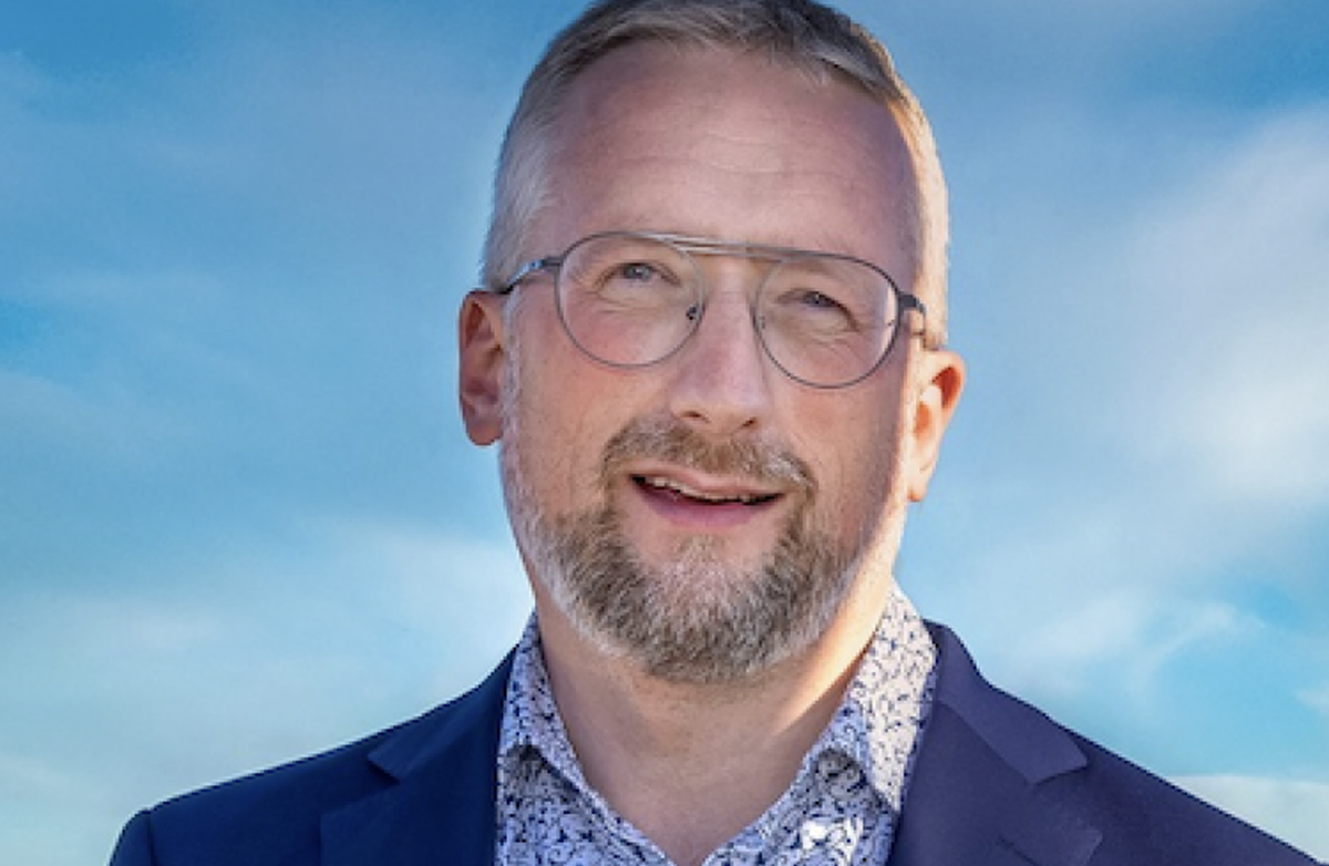 Middelaldrende mann i blå dress omgitt av blå himmel. Han smiler mot kameraet.