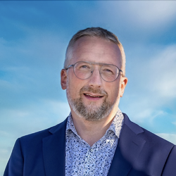 Middelaldrende mann i blå dress omgitt av blå himmel. Han smiler mot kameraet.